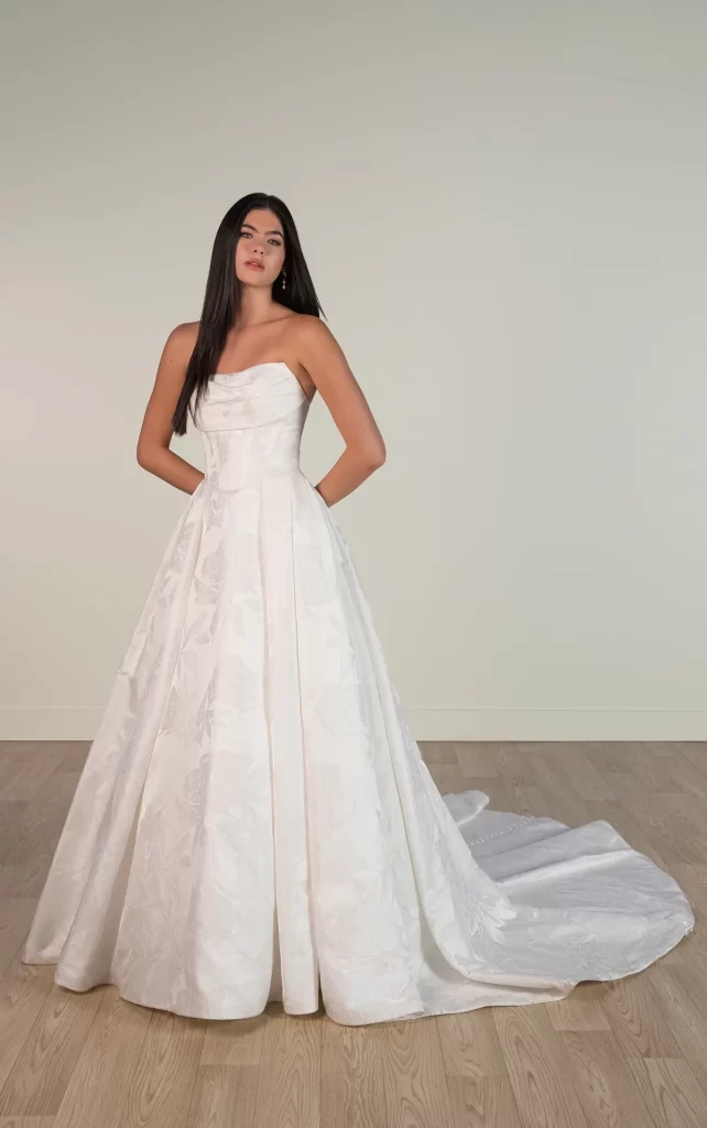 8084-Stella York-Pure Bridal. Edmonton bridal shop, Edmonton wedding dress. For the bride who blends modern sophistication with timeless romance, Style SY8084 brings together whimsical fairytale charm and contemporary designer fashion. This stunning strapless ballgown features a ruched neckline that wraps elegantly into a basque waist bodice. Contoured seams beautifully shape the figure before flowing into a pleated skirt, accented with a gown-length row of fabric-covered buttons for a classic touch. The pleated waist enhances the princess silhouette, which then expands into a voluminous ballgown skirt crafted from luxurious grand satin, completing the sleek fashion-forward look. This gown is also available in plus sizes.