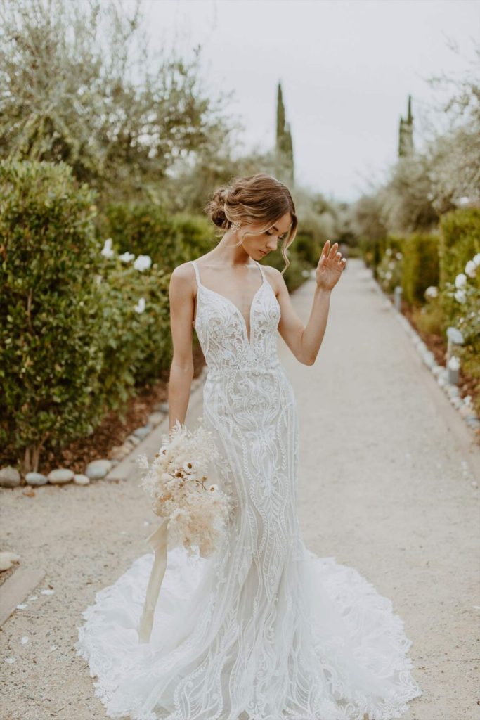 D3488-Essense of Australia-Pure Bridal-Lacy-Sexy-Modern-Edmonton-Bridal Shop-Wedding-Gown, Vintage glamour and modern luxe blend effortlessly in Style D3488. Swathed in beautifully beaded scrolling and linear lace, this dazzling gown accentuates the figure in a body-skimming fit-and-flare silhouette. Sparkling details adorn the graceful spaghetti straps before framing an ultra-flattering deep plunging neckline with illusion tulle. The sexy side cutouts lead to a daring V-shaped open back with artful linear beading and a fabric-covered button back detail. From the natural waist, the alluring vertical beaded graphics conclude in an oh-so-romantic lace-trimmed scalloped train.