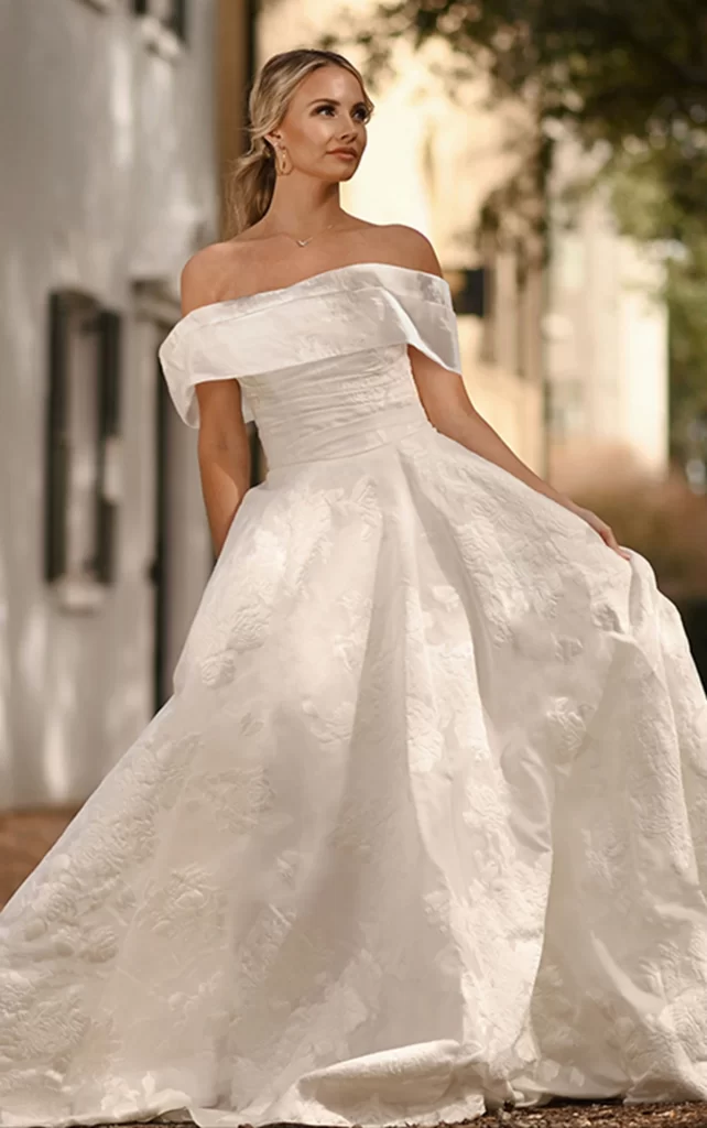 D3971-Essense of Australia-Pure Bridal. Edmonton bridal shop. Edmonton wedding dress. Exuding timeless elegance, Style D3971s luxe floral-print organdy jacquard and tailored fit emanate style and sophistication. The princess ballgown features a detachable off-the-shoulder shawl that fastens atop a strapless wrapped bodice fastened by a gown-length row of fabric-covered buttons. At the waist, a full ballgown skirt flatters the silhouette and highlights the unique printed organdy jacquard. Luxurious and modest, this elegant organdy jacquard ballgown makes a luxurious fashion statement down the aisle. This regal gown is also available in plus sizes.