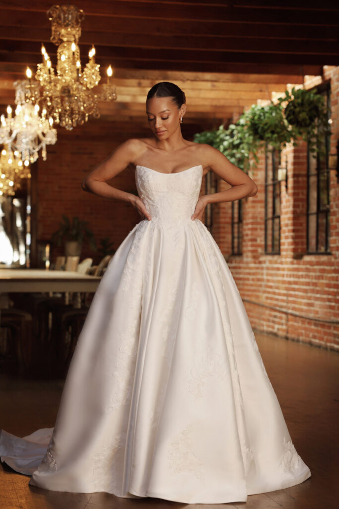 Pure Bridal-Essense of Australia-D4265-A modern take on classic bridal glamour, Style D4265 exudes timeless elegance with its striking ballgown silhouette and luxurious Mikado fabric. The strapless bodice showcases a chic scooped cat-eye neckline, with expertly placed seaming that highlights the natural hourglass shape. From the defined Basque waist, sculpted pleats unfold into a voluminous ballgown skirt, creating a breathtaking, regal effect. A row of fabric-covered buttons trails from the bodice to the hem of the dramatic train, adding a touch of refined sophistication. Designed for the fashion-forward bride, this clean and structured gown makes an unforgettable statement. This gown is also available in plus size.