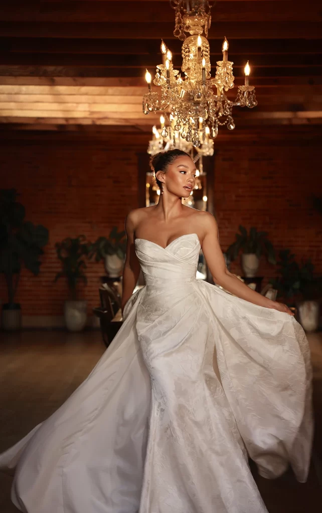 D4300-Essense of Australia-Pure Bridal. Edmonton bridal shop, Edmonton wedding dress. Timeless elegance meets modern sophistication in Style D4300, a stunning strapless column wedding dress designed for the bride who loves both classic and contemporary style. Crafted from luxurious satin jacquard, the structured bodice features a draped sweetheart neckline with delicate ruching that enhances the figure. The sleek column silhouette is complemented by a detachable pleated overskirt, adding dramatic movement and versatility. A row of fabric-covered buttons trails down the back for a vintage-inspired touch, blending refined glamour with effortless romance. This gown is also available in plus sizes.