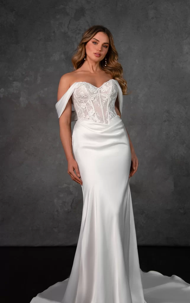 D4303-Essense of Australia-Pure Bridal. Edmonton bridal shop, Edmonton wedding dress. For the bride with a flair for modern glamour, this sleek charmeuse column wedding gown delivers effortless sophistication. The off-the-shoulder sweetheart neckline frames the structured, sheet lace corset bodice, adorned with intricate beading for a touch of sparkle. Flowing from the fitted bodice, the luxe charmeuse skirt drapes beautifully over the figure, creating a smooth, sultry silhouette with a row of fabric-covered buttons cascades down the back, leading to a soft, sweeping train for an elegant finish. Blending timeless romance with contemporary minimalism, this designer wedding dress is a showstopping choice for the fashion-forward bride.