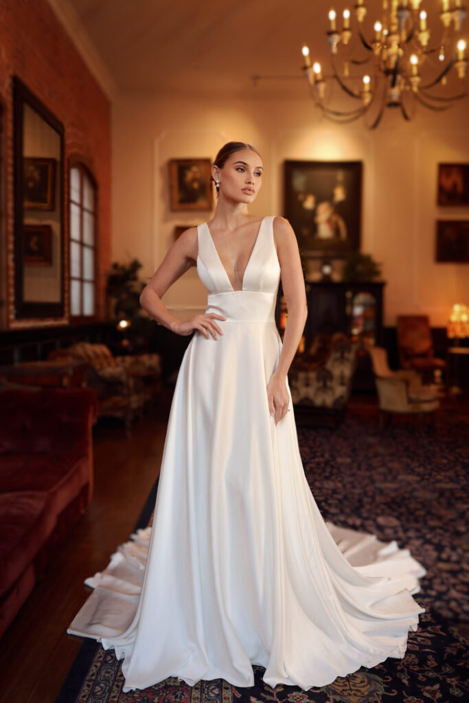 Pure Bridal-Essense of Australia-D4359-Edmonton Bridal Shop-Style D4359 is a sleek, modern satin wedding gown that brings high-fashion elegance to life. Featuring a bold deep V-neckline, this A-line silhouette captures attention with its sculpted bodice, creating a striking hourglass shape. Illusion tulle lining adds an element of modesty while maintaining comfort, allowing the daring design to shine. A matching V-back mirrors the neckline, further enhancing the gown's allure. From the empire waist, a flowing pleated skirt cascades gracefully, showcasing the luxurious satin fabric in its full, radiant beauty. This wedding gown is also available in plus sizes.