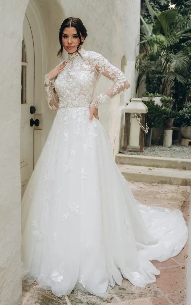SY8126–Stella York-Pure Bridal, Edmonton bridal shop, Edmonton wedding dress. Timeless bridal beauty with a modern twist, this breathtaking ballgown is a vision of romance. Intricate mixed floral lacework creates a unique, one-of-a-kind design, forming a high neckline and sheer bodice that blend classic elegance with contemporary allure. Cascading lace trails down the full skirt, crafted from layers of tulle and organza for a voluminous yet airy effect. At the back, a delicate keyhole detail framed by floating lace elements adds the finishing touch. Effortlessly blending tradition and modernity, this enchanting gown is also available in plus sizes and with a variety of bodice lining options.