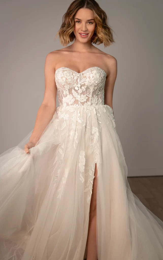 SY8139-Stella York-Pure Bridal, Edmonton bridal shop, Edmonton wedding dress. A captivating blend of romance and modern glamour, this strapless wedding dress is designed to make an unforgettable statement. Style SY8139 shimmers with sequin floral lace, gracefully cascading over a soft A-line silhouette for an effortlessly elegant look. The sheer bodice, adorned with intricate floral embellishments, features a sweetheart neckline and exposed corset boning, striking a balance between sultry and sophisticated. From the waist, airy layers of tulle create a dreamy, flowing skirt, while a high leg slit adds a bold, fashion-forward touch. Designed for the bride who loves timeless beauty with a modern edge. This gown is also available in plus sizes.